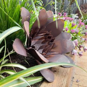 Rusted metal flower sculpture made from Corten steel, featuring a rich orange-red patina, set in a garden. The sculpture measures 36cm in diameter and adds rustic charm to outdoor spaces.